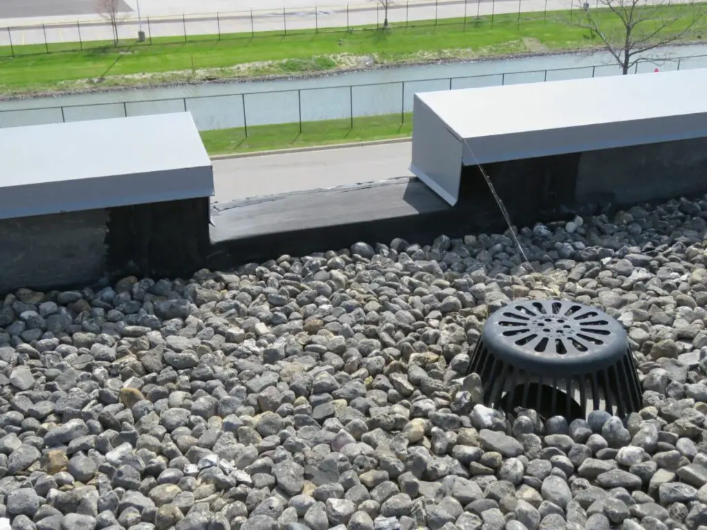 A channel-type roof scupper next to a roof drain on a commercial flat roof.