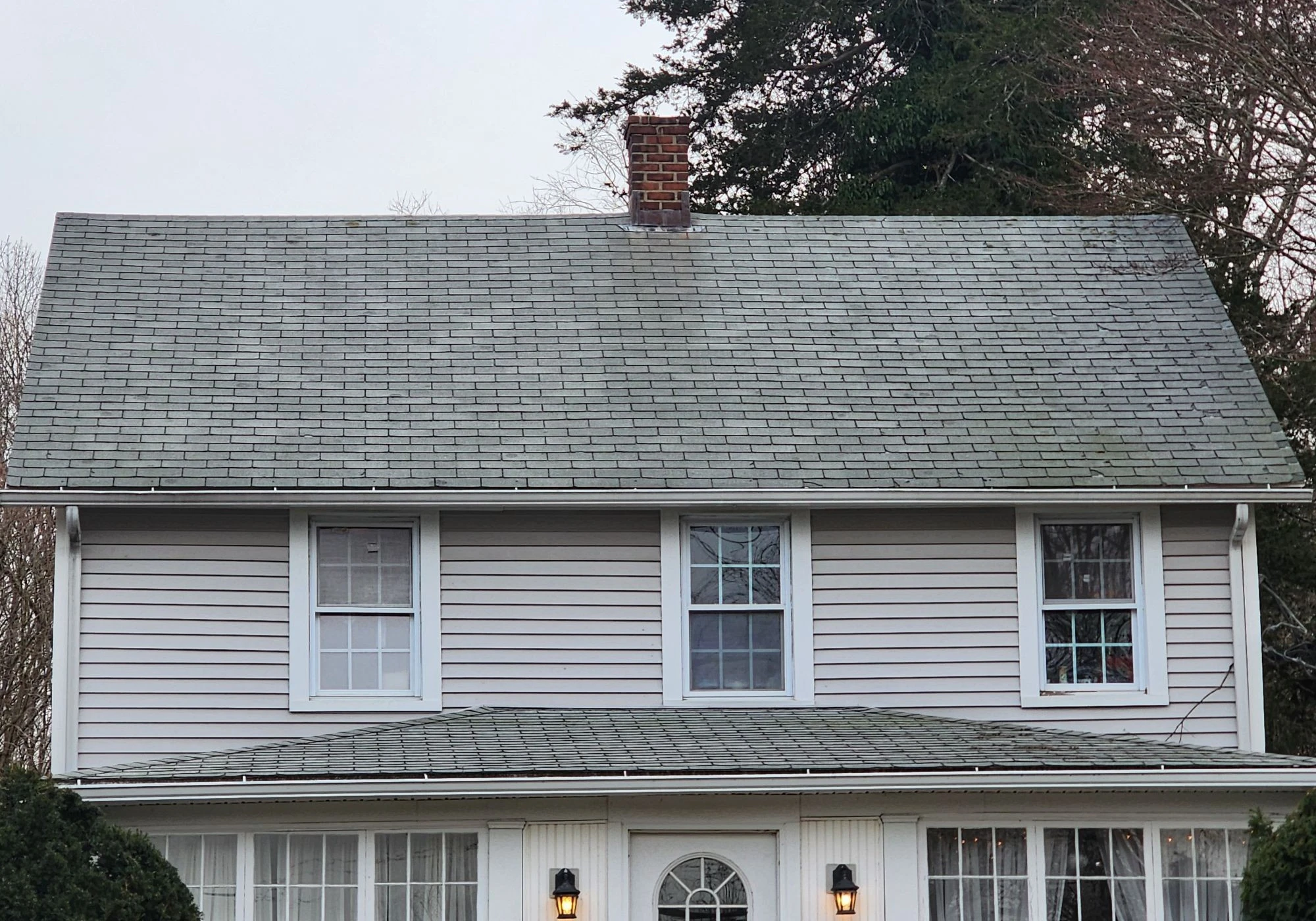 A house with an asphalt shingle roof with a chimney in the middle and a lower one-story roof.